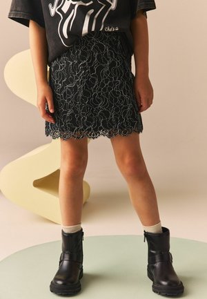 Zwarte kanten rok met een gekartelde zoom, ingewikkeld bloemenpatroon, gedragen met kniehoge zwarte laarzen en crèmekleurige sokken. Grijze oversized t-shirt erboven.
