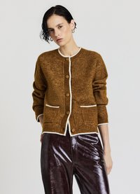 Braune Strickjacke mit weißem Rand, rundem Ausschnitt und Knopfverschluss. Mit zwei aufgesetzten Taschen vorne und langen Ärmeln. Glänzende braune Hose.