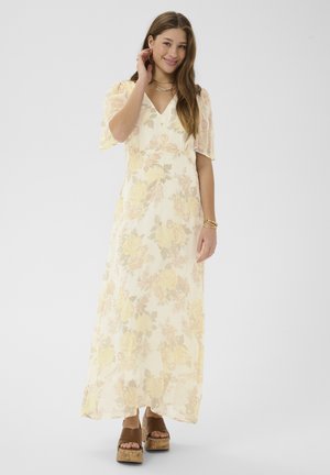 Kvinde iført en lang, lys blomsterkjole og brune platåsandaler, smilende med den ene hånd, der rører ved hendes hår.