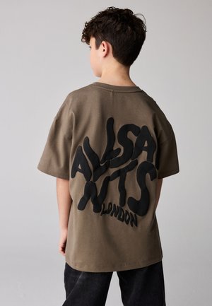 Junge mit kurzen lockigen Haaren, trägt ein olivgrünes T-Shirt mit großem schwarzem Schriftzug "AllSaints London" auf dem Rücken, steht mit dem Rücken zur Kamera.