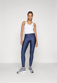 Hvit atletisk tank topp og marineblå leggings med en jevn tekstur. Modellen har også på seg grå joggesko med et mønstrete design.
