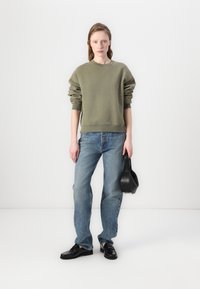 Olijfgroen oversized sweatshirt met geribbelde manchet, gecombineerd met lichte denim jeans. Een zwarte handtas en glanzende zwarte schoenen maken de outfit compleet.