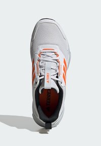 Grijze sportieve schoen met oranje accenten, bovenwerk van textuurstof, versterkte neuskap en zwarte binnenvoering. Voorzien van een trekband aan de hiel.