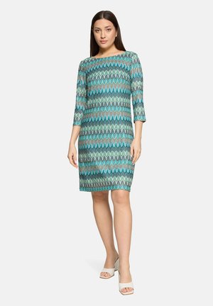 Kvinde stående iført en knælængde, langærmet kjole med turkis, blå og grønne mønstre samt hvide åbne tå-sandaler mod en ensfarvet baggrund.