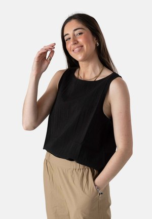 Joven sonriente, con una camiseta negra sin mangas, pantalones beige, collar de oro y pulseras, una mano levantada cerca de la cara, la otra en el bolsillo.