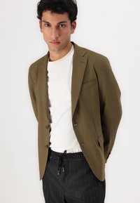 Blazer verde oliva con dos botones, solapas con muesca y un bolsillo en el pecho. Combinado con una camiseta blanca y pantalones de rayas negras.