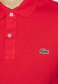 Červené polo tričko vyrobené z texturované tkaniny, s klasickým límcem a třemi bílými knoflíky. Obsahuje zelenou vyšívanou logo krokodýla.