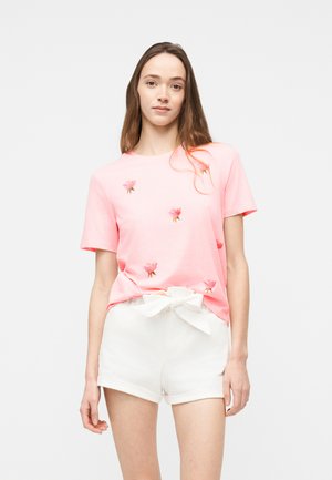 Jonge vrouw die een roze T-shirt met bloemrijke borduurwerk en witte korte broek draagt, met een voorstrik, staand tegen een effen achtergrond.