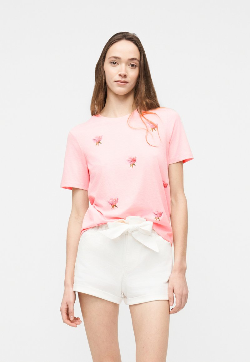 Νεαρή γυναίκα που φοράει ροζ μπλουζάκι με κεντήματα λουλουδιών και λευκές σορτς δεμένες με φιόγκο μπροστά, στέκεται μπροστά από ένα απλό φόντο.