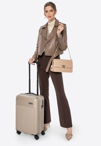 Beige Hartschalenkoffer mit horizontaler Rillenstruktur, ausziehbarem Griff und vier Rollen. Das Modell trägt eine braune Lederjacke und beige Accessoires.