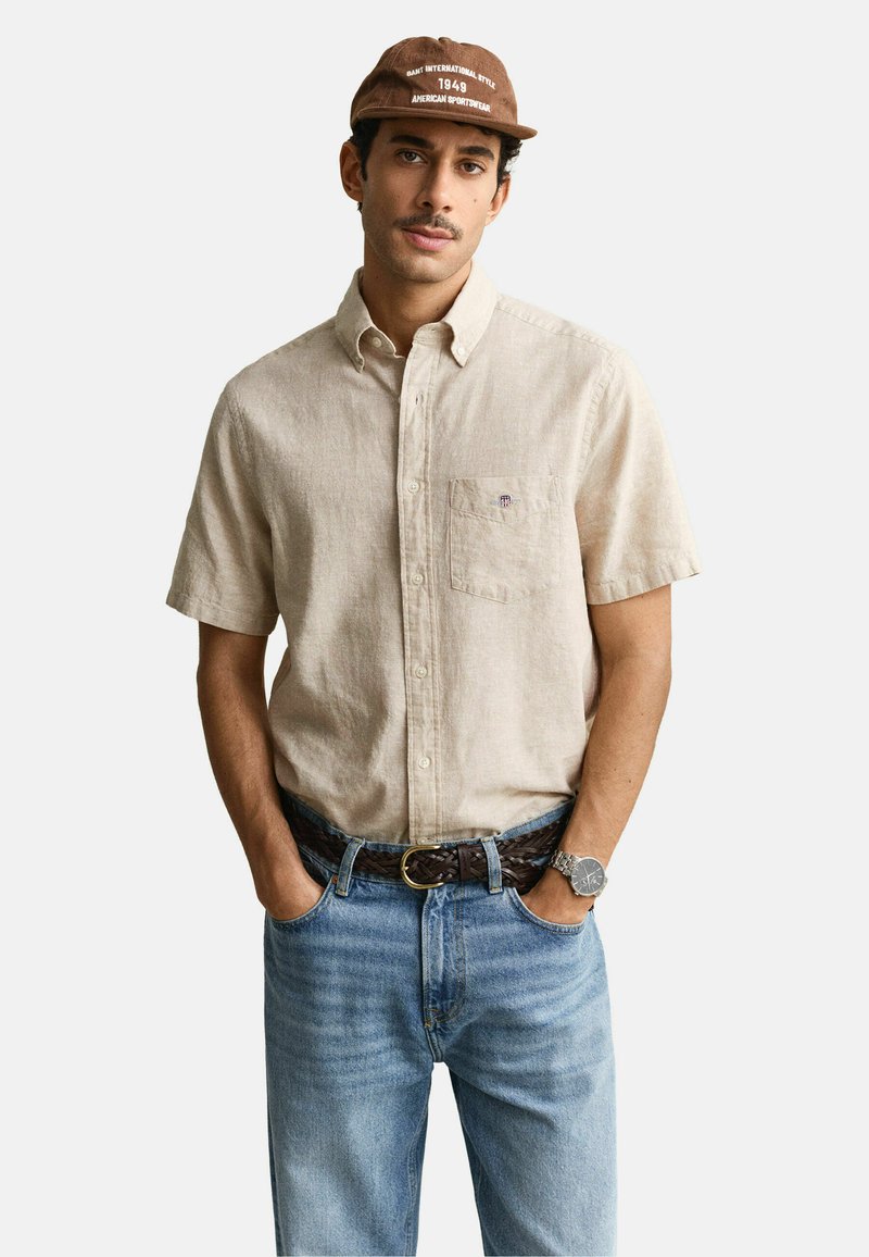 Homme portant une chemise beige à manches courtes avec boutons, un jean bleu clair, une ceinture tressée marron, une casquette marron et une montre-bracelet, mains dans les poches.