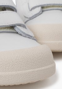 Zapatos de lona blancos con correas de velcro, suelas de goma texturizada en beige y detalles en azul claro a lo largo de las correas. Vista en primer plano de los materiales.