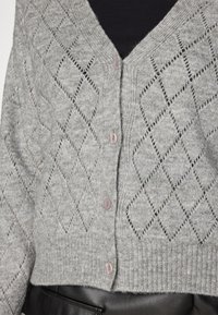 Light gray knitted cardigan with a diamond pattern, featuring a deep V-neck and five buttons. Ribbed hem and soft texture.
