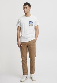 White short-sleeve T-shirt with a palm-patterned pocket, paired with light brown cargo pants and white sneakers. Simple, casual design.