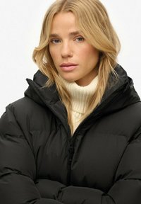 Femme blonde avec des cheveux lâches portant un pull à col montant blanc et une doudoune noire zippée, regardant légèrement sur le côté.