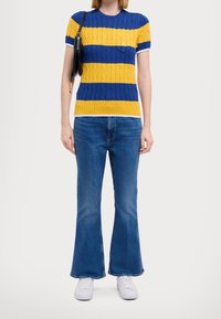 Knit short-sleeve sweater with blue and yellow horizontal stripes, featuring a cable pattern. Paired with flared blue jeans and white sneakers.