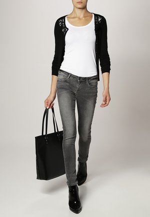 Žena v černém kardiganu, bílém tílku, šedých úzkých džínách a černých kotníkových botách, nesoucí velkou černou tašku typu tote.