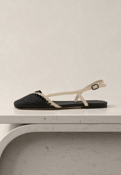 Black woven flat sandal with beige braided straps and ankle buckle, displayed on a white textured surface against a neutral background.