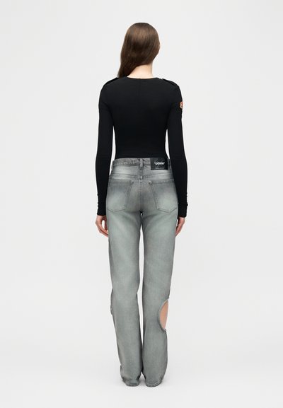 Femme aux longs cheveux bruns portant un haut noir à manches longues et un jean gris à jambes larges avec des découpes circulaires près des chevilles, debout devant un fond uni.