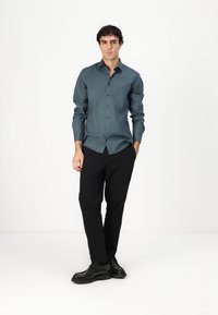 Chemise bleu canard à manches longues, assortie à un pantalon noir et des chaussures noires. La chemise présente une texture lisse et une coupe ajustée.