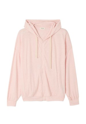 Femme portant un sweat à capuche rose clair zippé, un short en jean clair longueur genou, et des tongs noires à plateforme, debout devant un fond blanc uni.