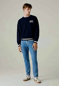 Marine sweatshirt med hvidt logo, ribbet manschetter og kant; parret med lyseblå jeans. Modellen bærer hvide sneakers med sorte detaljer.