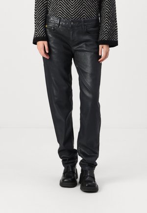 Personne portant un pantalon en cuir noir, des chaussures noires épaisses et un pull à motif zigzag noir et blanc, se tenant devant un fond blanc.