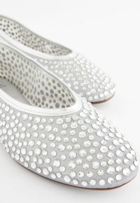 Chaussures plates blanches couvertes de strass clairs espacés régulièrement et doublure intérieure à pois sur fond blanc.