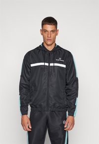 Chaqueta negra con cremallera y capucha, con detalles en blanco y verde azulado, puños elásticos y un logo en el pecho. Tela suave con un diseño deportivo.