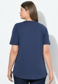 Žena s rovnými hnědými vlasy, oblečená v jednobarevném námořnickém modrém tričku s krátkým rukávem, čelí bílé zdi a má zády k fotoaparátu.