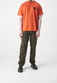 Persona de pie con una camisa de manga corta naranja, pantalones verde oliva y botas negras con cordones frente a un fondo blanco liso.