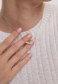 Srebrny naszyjnik z łańcuszka ozdobiony dwiema białymi perłami i trzema przezroczystymi koralikami, noszony na tle fakturowanego białego swetra. Ręka uniesiona nad naszyjnikiem.