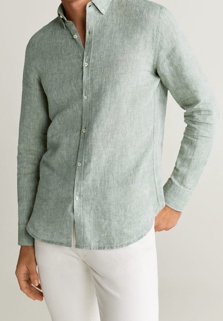 Homme portant une chemise en lin vert clair à manches longues avec des boutons blancs et un pantalon blanc, main partiellement dans la poche.