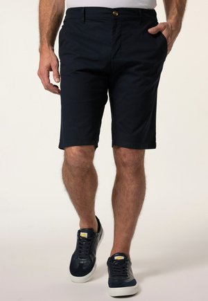 Mann trägt dunkelblaue Shorts und schwarze Sneakers mit weißen Sohlen, steht mit einer Hand in der Tasche.