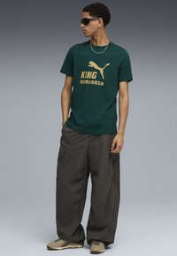 Mørkegrøn bomulds T-shirt med gylden "KING BORUSSIA" tekst og Puma-logo; parret med løstsiddende grå cargo bukser og tan sneakers.
