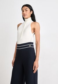 Αμάνικο κρεμ τοπ με ψηλό γιακά και μπροστινό φερμουάρ, συνδυασμένο με φαρδιά ναυτικά παντελόνια με λευκές οριζόντιες ρίγες και χρυσή λεπτομέρεια.