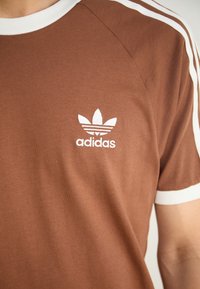 Ruskea puuvillainen t-paita, jossa on valkoiset olkaiterät. Takana on valkoisella brodeerattu Adidas-logo rinnassa, pyöreä kaula-aukko.