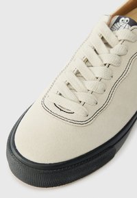 Gai bēšs zamša sporta apavi ar teksturētu virsmu, melnu gumijas zoli un kontrastējošu tumšu šuvumu. Iespējas: plakani bēši šņores un logo uz mēles.