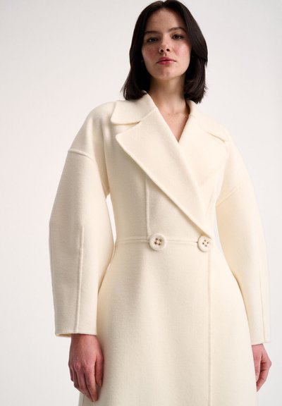 Double-breasted wool coat in cream with wide lapels, oversized long sleeves, and two decorative buttons at the waist. Smooth texture.