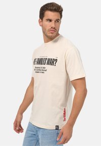 Camiseta de algodón beige con texto negro en negrita en la parte delantera, mangas cortas y texto rojo en el dobladillo lateral. Ajuste relajado, textura suave.