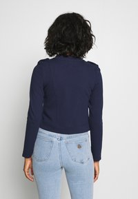 Marinblå kort jacka med långa ärmar och axelaccenter. Enkelt mönster med en slät yta, kombinerad med ljusblå högmidjade jeans.