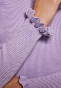 Maglione lilla a coste con polsini arricciati; presenta una texture morbida e un design aderente che mette in risalto l'area del braccio e del polso.