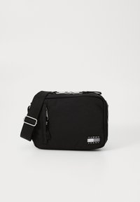 Sort nylon crossbody taske med lynlåslukning, justerbar rem og et logo-patch. Rektangulær form, glat tekstur.