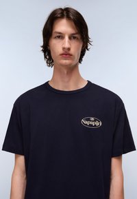 Navy blue cotton t-shirt with a crew neck. Features a gold logo patch on the left side of the chest. Short sleeves, relaxed fit.