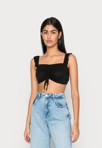 Top cropped preto com alças de ombro plissadas, detalhe de laço frontal e um acabamento texturizado, combinado com jeans de cintura alta azul claro.