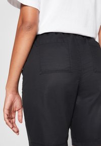 Vue arrière d’une personne portant un short noir avec des poches plaquées et une chemise blanche rentrée, main détendue le long du corps.