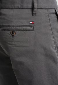 Zbliżenie na kieszeń szarych spodni z zapięciem na guzik oraz małym prostokątnym detalem z materiału w kolorach czerwonym, białym i niebieskim nad kieszenią.