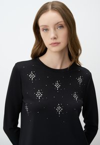Maglietta nera a maniche lunghe con scollo rotondo, decorata con gemme argentate in motivi floreali e a stella sulla parte anteriore. Tessuto morbido.