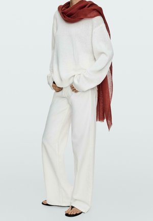 Persona che indossa un maglione di lana bianco, pantaloni bianchi larghi, infradito nere e una grande sciarpa rosso ruggine drappeggiata su una spalla.