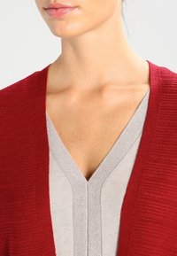 Czerwony sweter kardigan o fakturze, noszony na jasnoszarym topie z dekoltem w serek. Kardigan ma prążkowany wzór i otwarty przód.